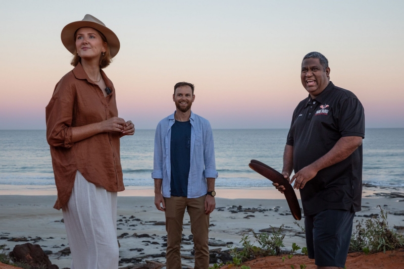 An Aboriginal guide holding traditional clapsticks speaks with two tourists on the shoreline at sunset during a Mabu Buru Tours experience at Cable Beach in Broome, Western Australia, with quiet ocean waves breaking behind them. © Tourism Australia