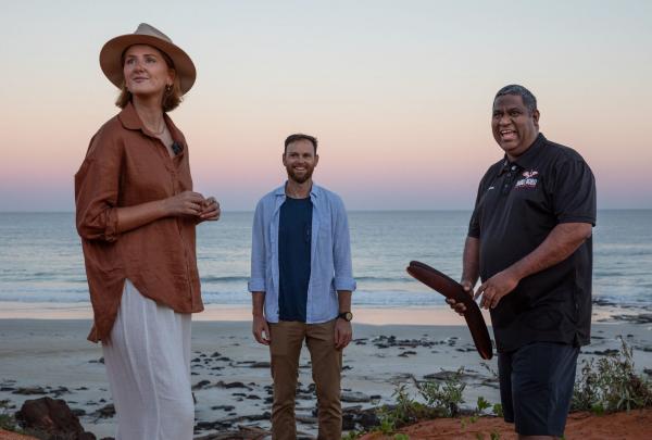 An Aboriginal guide holding traditional clapsticks speaks with two tourists on the shoreline at sunset during a Mabu Buru Tours experience at Cable Beach in Broome, Western Australia, with quiet ocean waves breaking behind them. © Tourism Australia