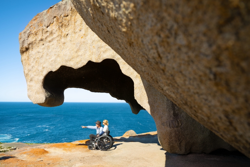 Amidst the vibrant hues of Kangaroo Island's Remarkable Rocks, a person in a wheelchair and their enthusiastic tour guide share a moment of awe. Positioned against the brilliant, multicoloured rocks, they gaze out over the azure expanse of the Southern Ocean. The dynamic contrast between the vivid geological formations and the deep blue sea creates a visually stunning panorama, echoing the accessibility and inclusivity of the natural wonder they're enjoying © Tourism Australia