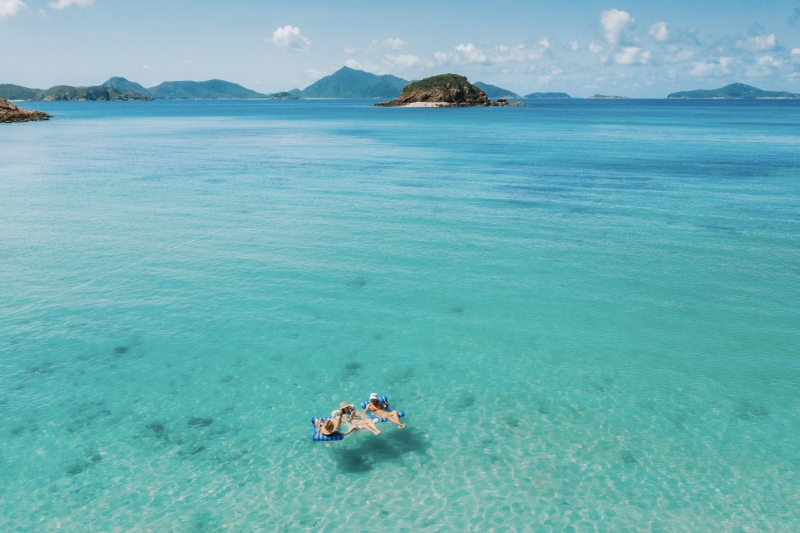 Three individuals recline on floating lounges, basking in the radiant sunshine while immersed in the crystal-clear turquoise waters of Whitehaven Beach on Whitsunday Island. The stunning scene epitomises a perfect day in paradise.. © Tourism Australia