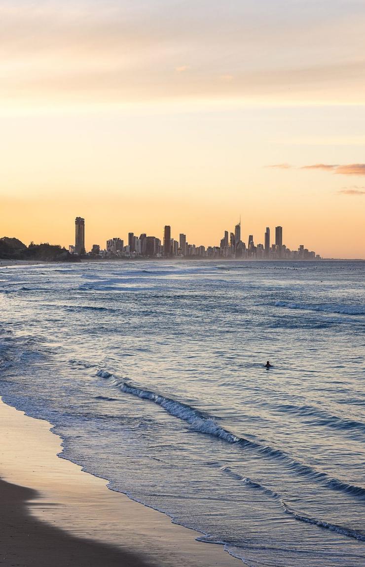 As the sun sets, the sky glows with a warm blend of oranges and pinks, casting a serene light over the lapping waves of Burleigh Heads on the Gold Coast, Queensland © Tourism Australia