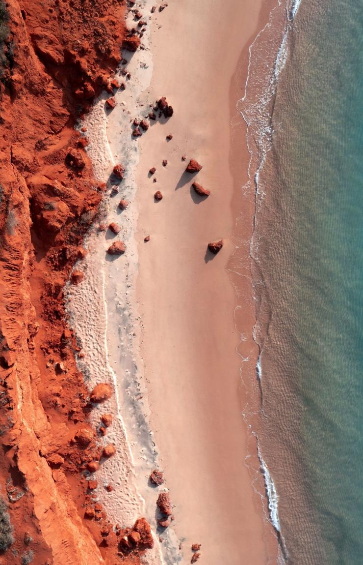 Aerial view of striking red dirt cliffs meeting sandy beach and turquoise clear ocean waters at Francois Peron National Park, Shark Bay, where vibrant coastal landscape creates vivid colour contrasts. © Tourism Australia
