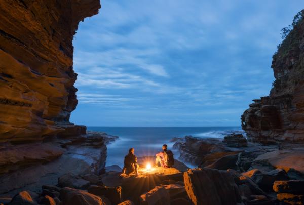 Two people sitting on a rock at night looking out to the ocean at Avoca Caves, New South Wales © Tourism Australia