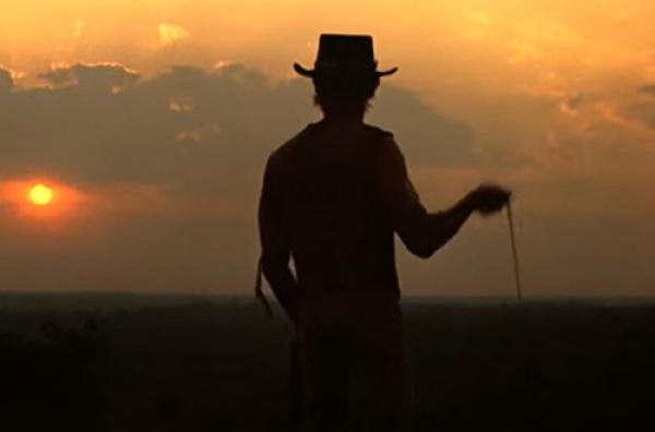 A shadowed silhouette of a man in an outback hat stands against a vibrant orange sunset