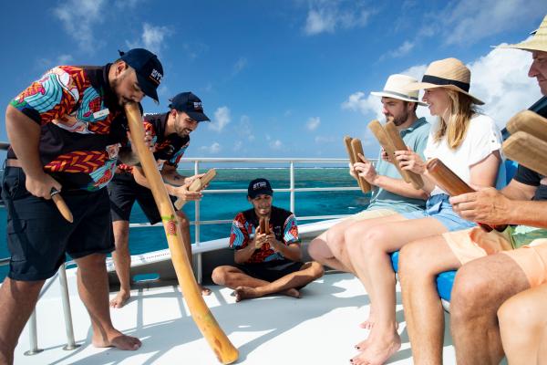 On the sunny boat deck of Dreamtime Dive and Snorkel in Cairns, Queensland, Indigenous Sea Rangers play the didgeridoo and demonstrate clapsticks for a group of seated tourists holding their own wooden instruments.