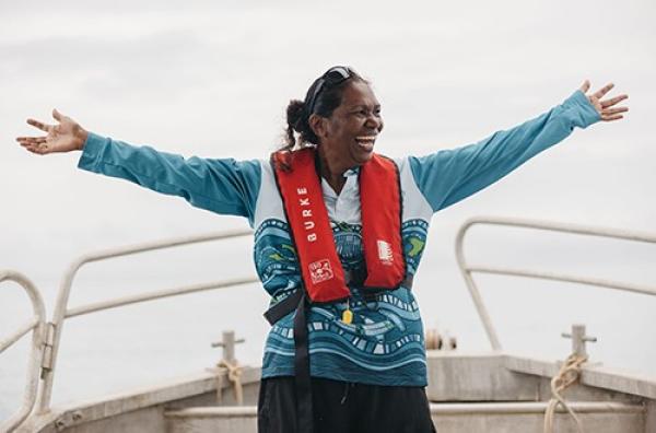 A guide on an Oolin Sunday Island Cultural Tour stands smiling with arms outstretched on bow of a boat.