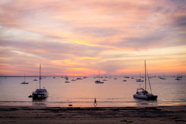 A silhouette of a person walking a dog along a wet beach at sunset, with catamarans moored in tranquil waters under a colorful sky