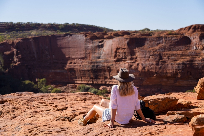 A lady, with a wide brimmed hat sitting with her back to the viewer, leaning back on her hands on a rich red rock platform, looking across a beautiful red escarpment of Canyon Rim Walk with Kings Canyon Resort, Kings Canyon, Central Australia, Northern Territory © Tourism Australia