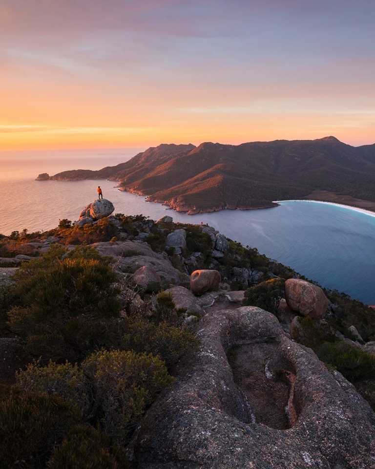 A hiker stands atop Mt Amos taking in the view of Wineglass Bay, Freycinet, Tasmania © Tourism Australia