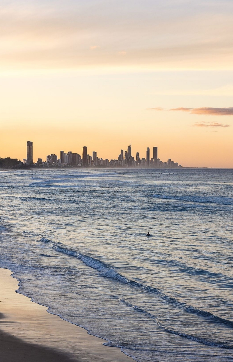As the sun sets, the sky glows with a warm blend of oranges and pinks, casting a serene light over the lapping waves of Burleigh Heads on the Gold Coast, Queensland © Tourism Australia