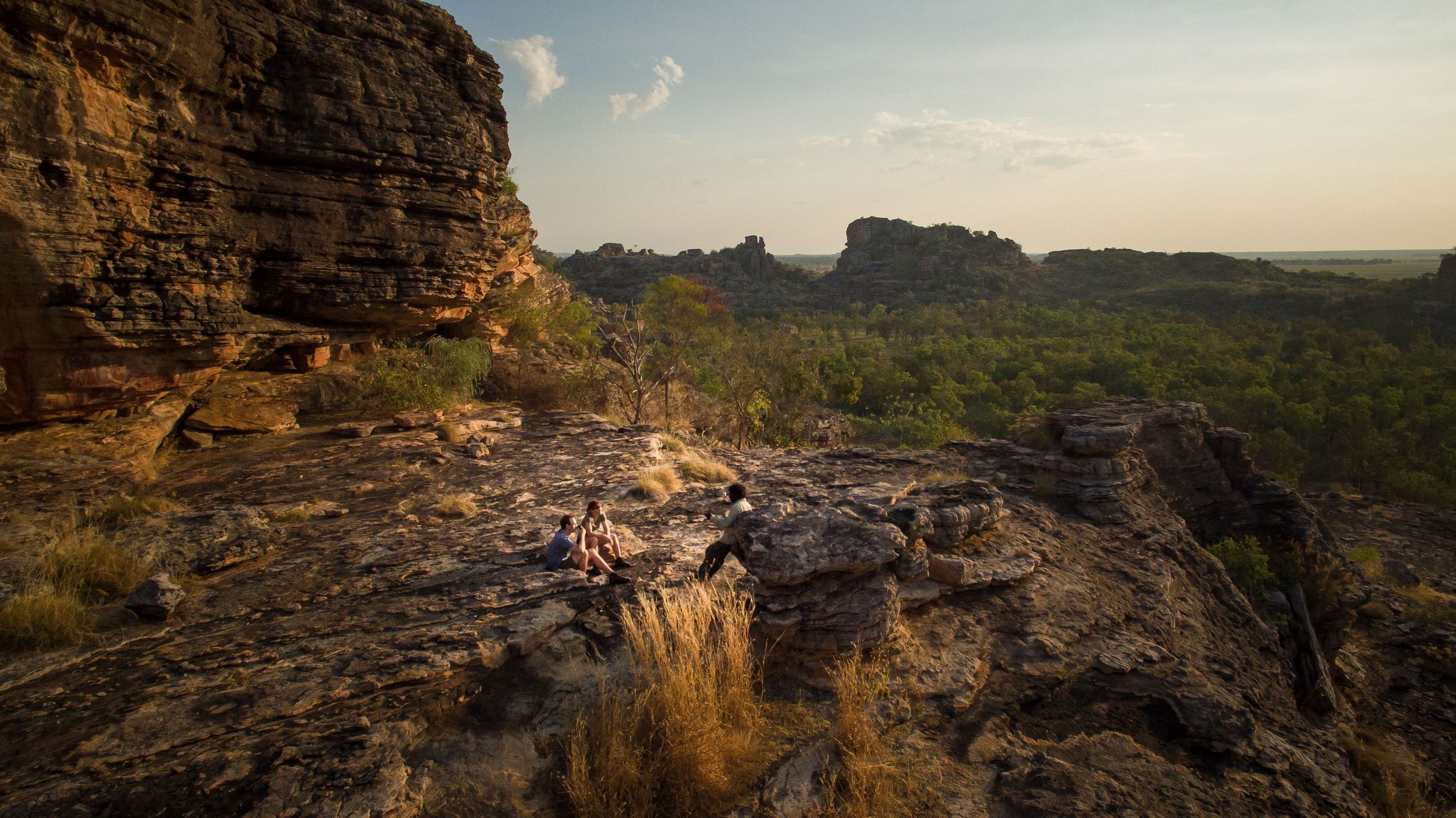 A tour guide talks with two visitors sitting on a rocky ledge overlooking the vast forested valleys and massive sandstone cliffs of Kakadu National Park, Northern Territory, during a Kakadu Cultural Tours excursion. © Tourism Australia